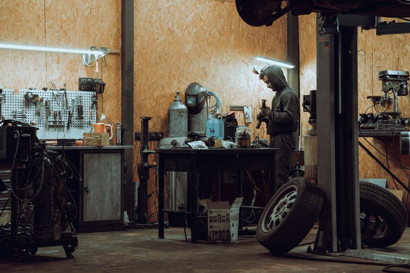 Mechanic working at a welding bench in a vehicle repair workshop, with a car raised on a hydraulic lift and a wheel removed in the foreground