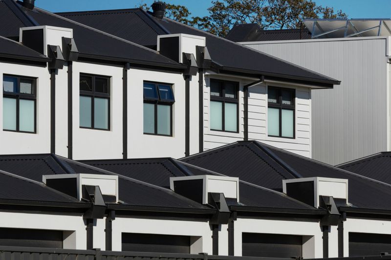 Rows of white townhouse rooftops with dark corrugated roofing, gable dormers and upper-storey windows in a multi-dwelling residential development